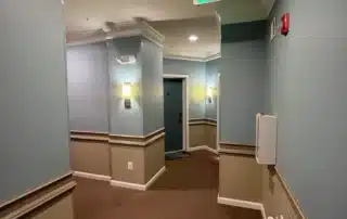 Hotel hallway with blue walls, tan wainscoting, brown carpet, and a green exit sign