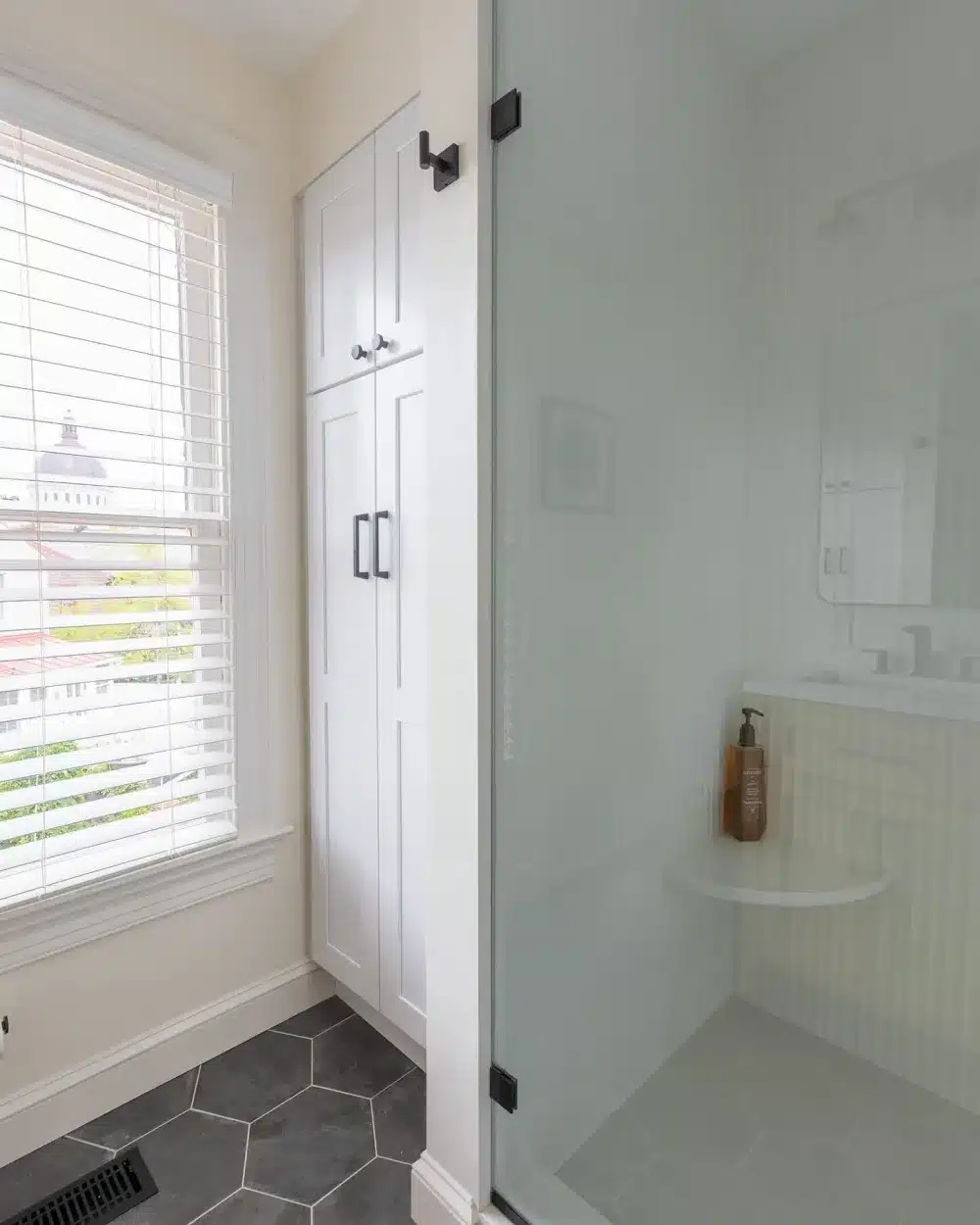 Custom white tall bathroom cabinetry next to a glass shower enclosure with dark hexagonal floor tiles
