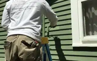 A painter in a white shirt and tan pants standing on a blue ladder painting green siding