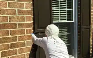 Painter in white uniform rolling dark brown paint onto a window shutter