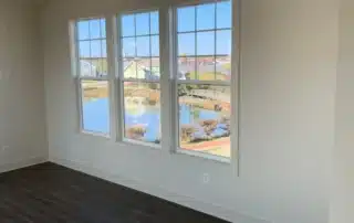 Empty room with dark hardwood floors, white walls, and a large triple window looking out at a pond