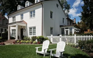 White two-story residential home with a green lawn and white Adirondack chairs