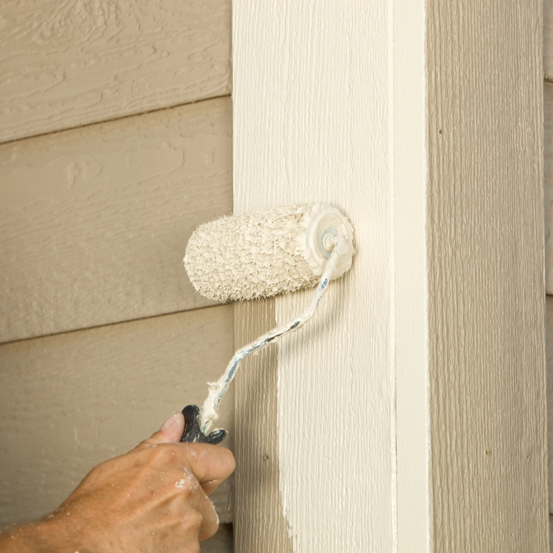 Painter using a roller to apply beige paint to a wooden house trim